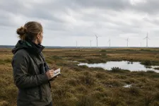 En ekolog med kikare och anteckningsblock i ett hedlandskap med vindkraftverk vid horisonten